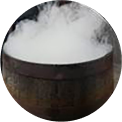 A circular image of a wooden barrel with thick white smoke or vapor billowing out from the top, creating a mysterious and dramatic effect.