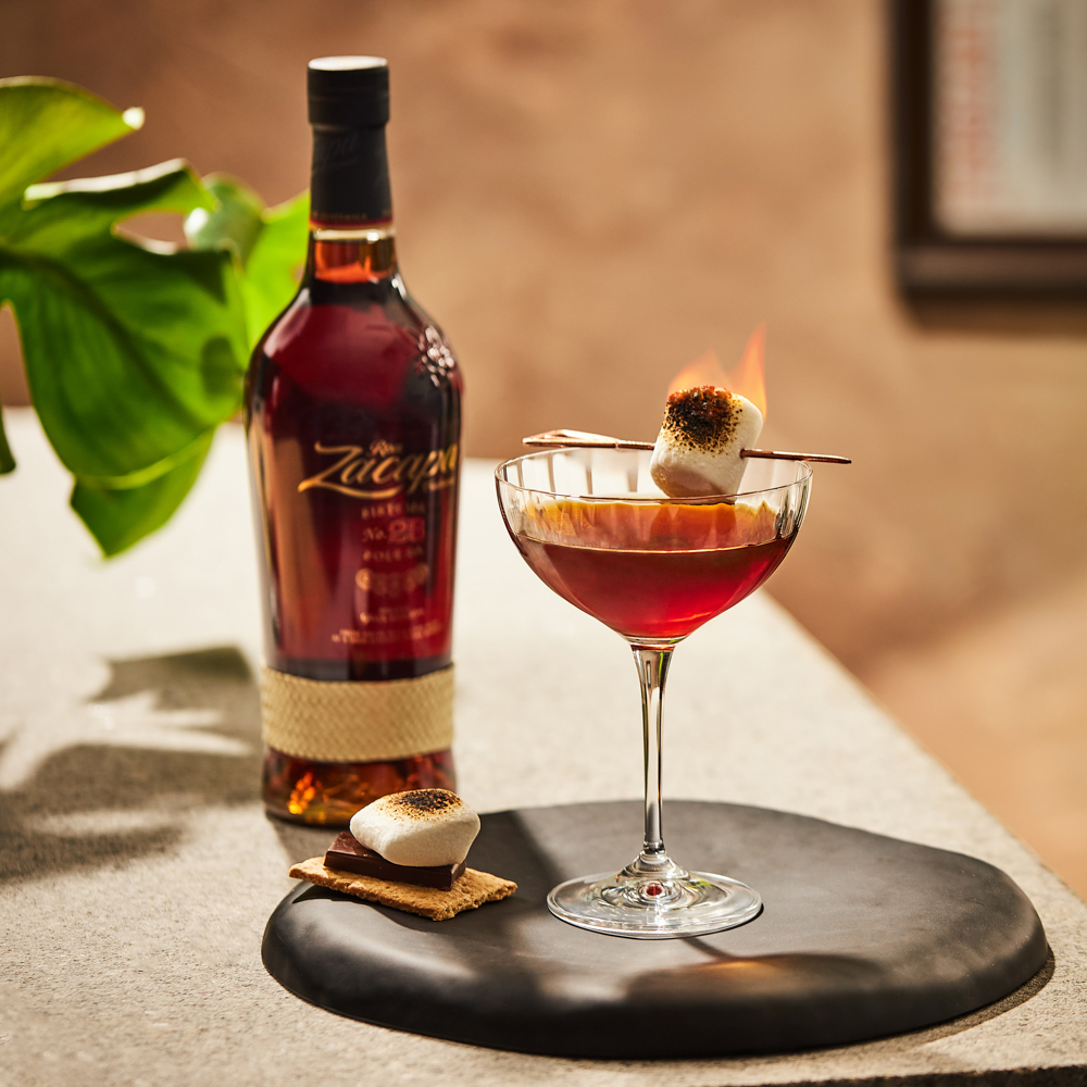 A bottle of Ron Zacapa rum stands next to a coupe glass filled with a rich, amber cocktail.