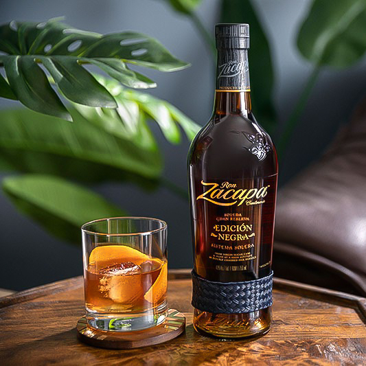 A bottle of Ron Zacapa Edición Negra rum is placed on a wooden table next to a glass containing a cocktail with a large ice cube and an orange peel garnish. 
