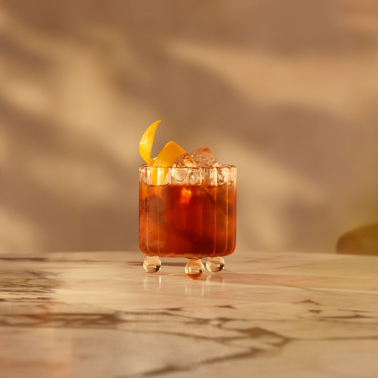 A glass with a unique rounded base sits on a marble surface, filled with an amber-hued cocktail and ice cubes.
