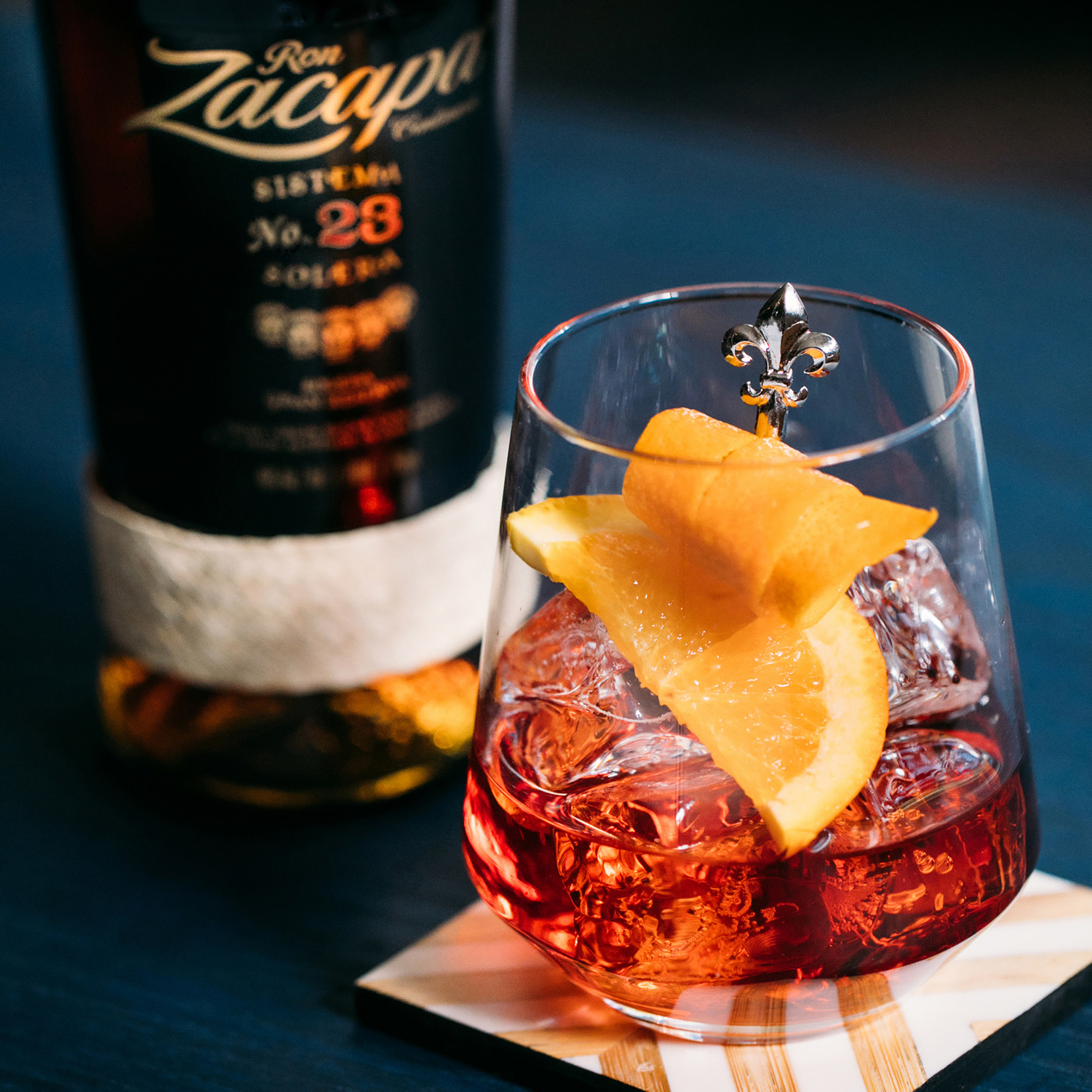 A glass of Ron Zacapa rum cocktail served over ice, garnished with a twist of orange peel and a fleur-de-lis cocktail pick.