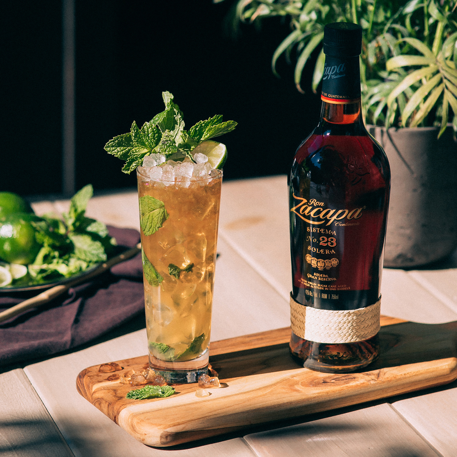 A bottle of Ron Zacapa Sistema 23 Solera rum is placed on a wooden serving board next to a tall glass filled with a refreshing mint garnish cocktail and crushed ice.