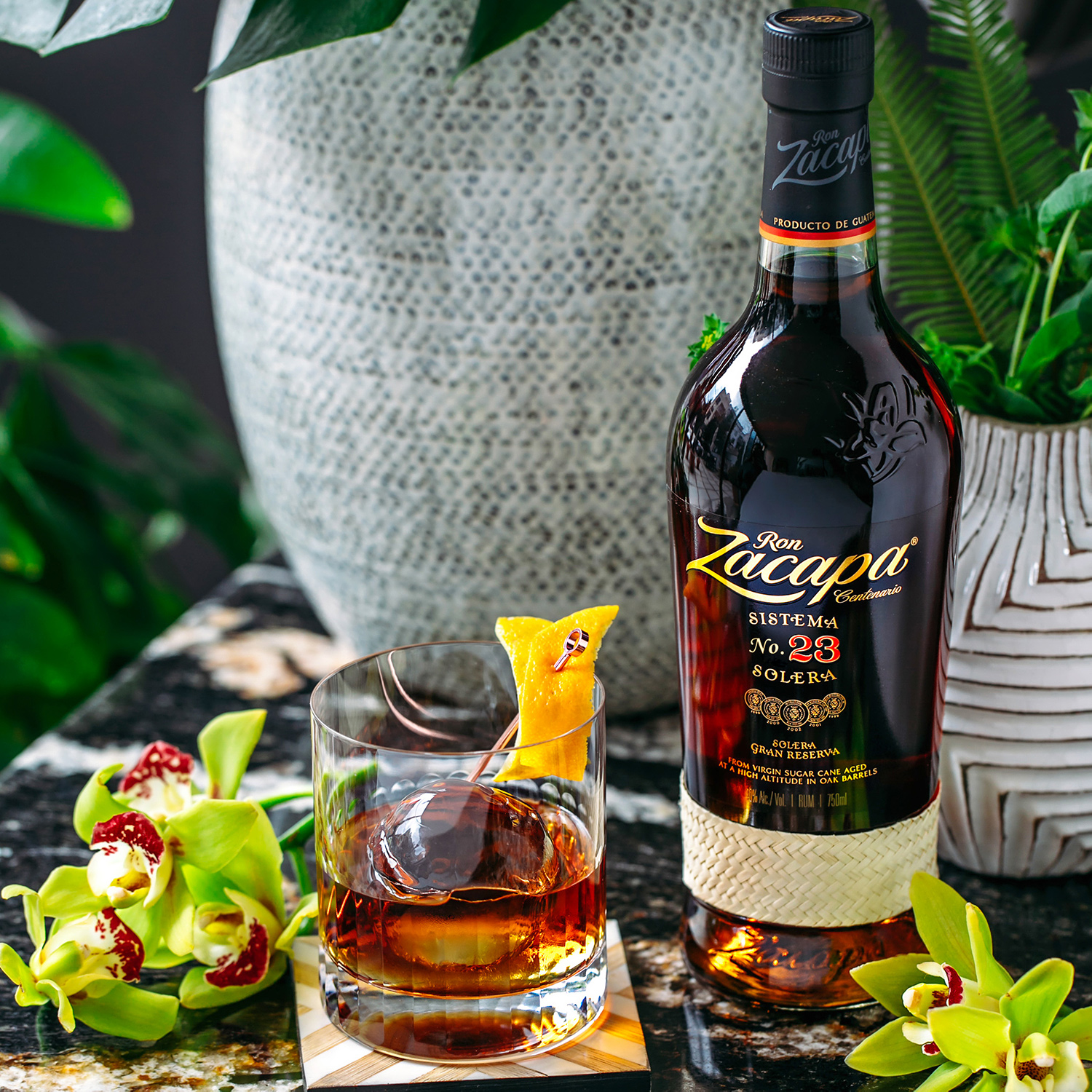 A bottle of Ron Zacapa Sistema 23 Solera rum stands on a marble surface next to a glass filled with a dark cocktail, garnished with an orange peel and served over a large ice sphere.