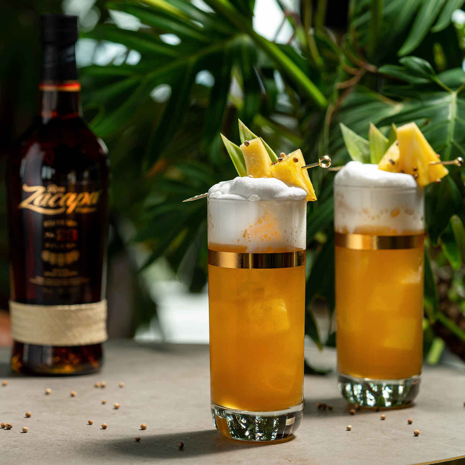 Two tall glasses filled with a frothy, golden cocktail are garnished with skewered pineapple wedges and leaves.