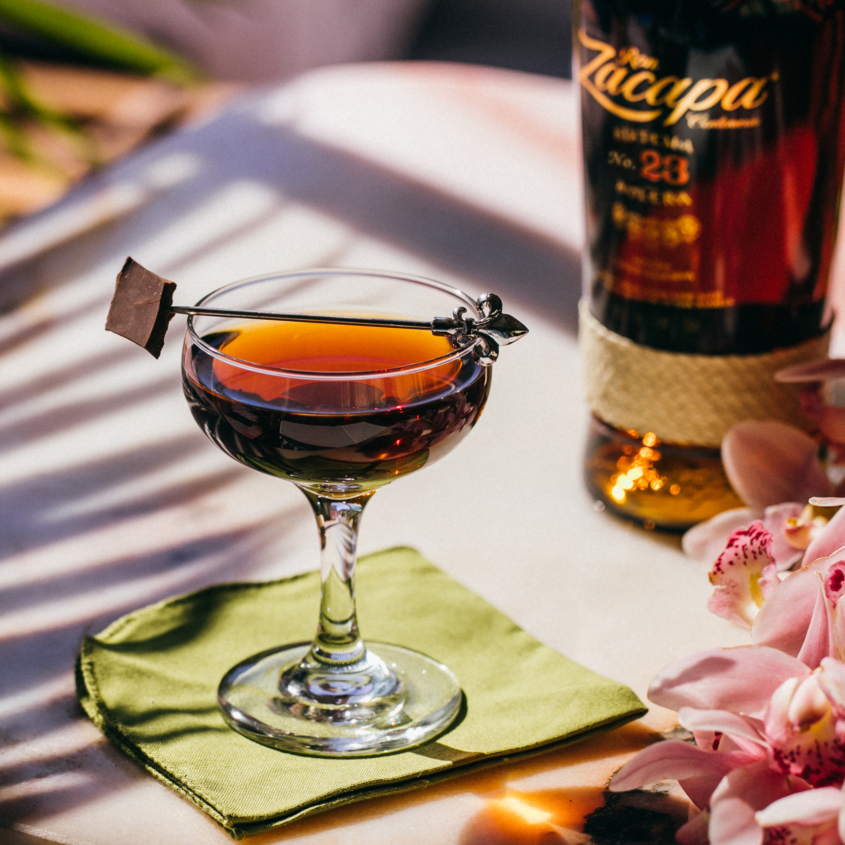 A cocktail glass filled with Ron Zacapa rum, garnished with a square piece of chocolate on a decorative metal pick.