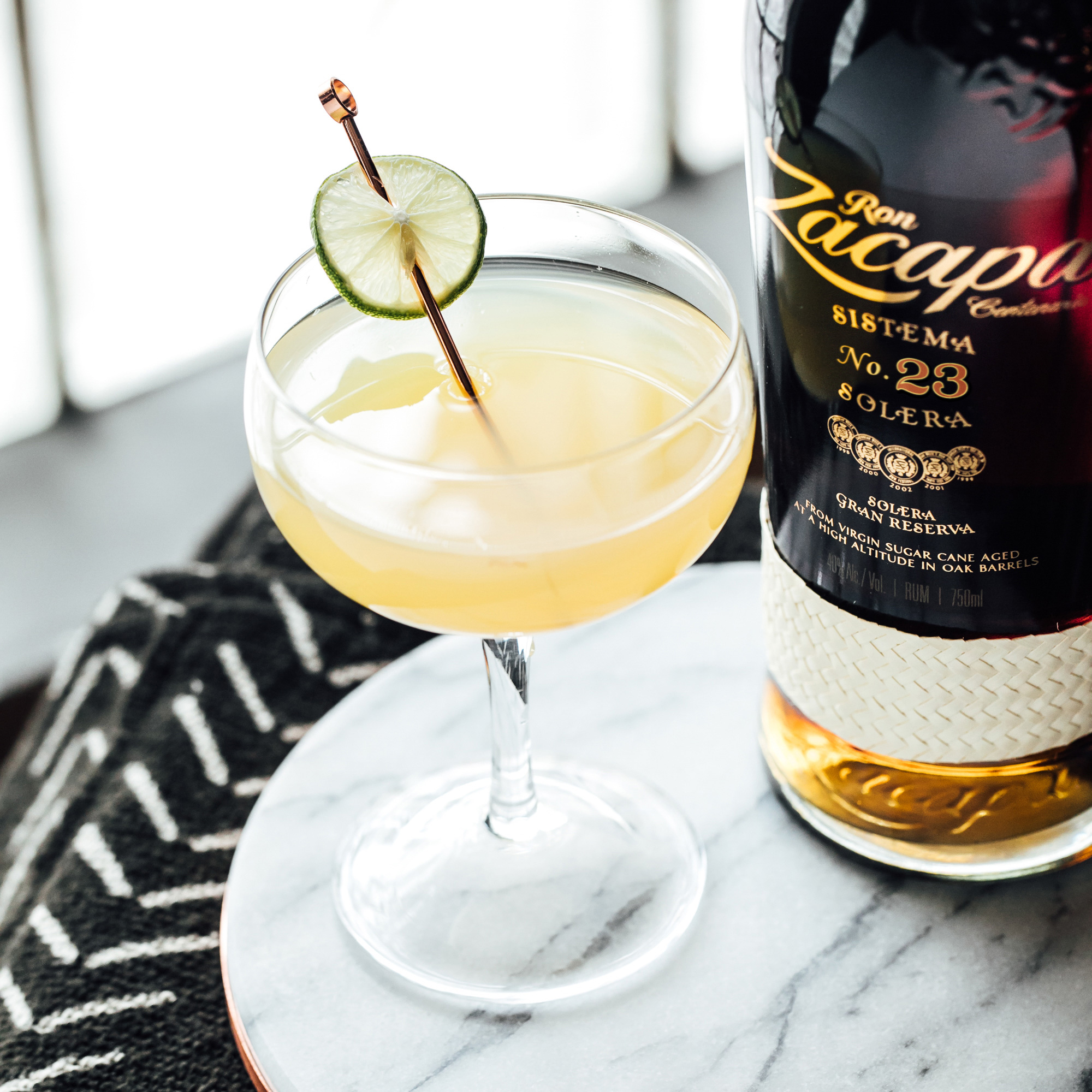 A coupe glass filled with a Ron Zacapa rum cocktail, garnished with a slice of lime on a metal pick.