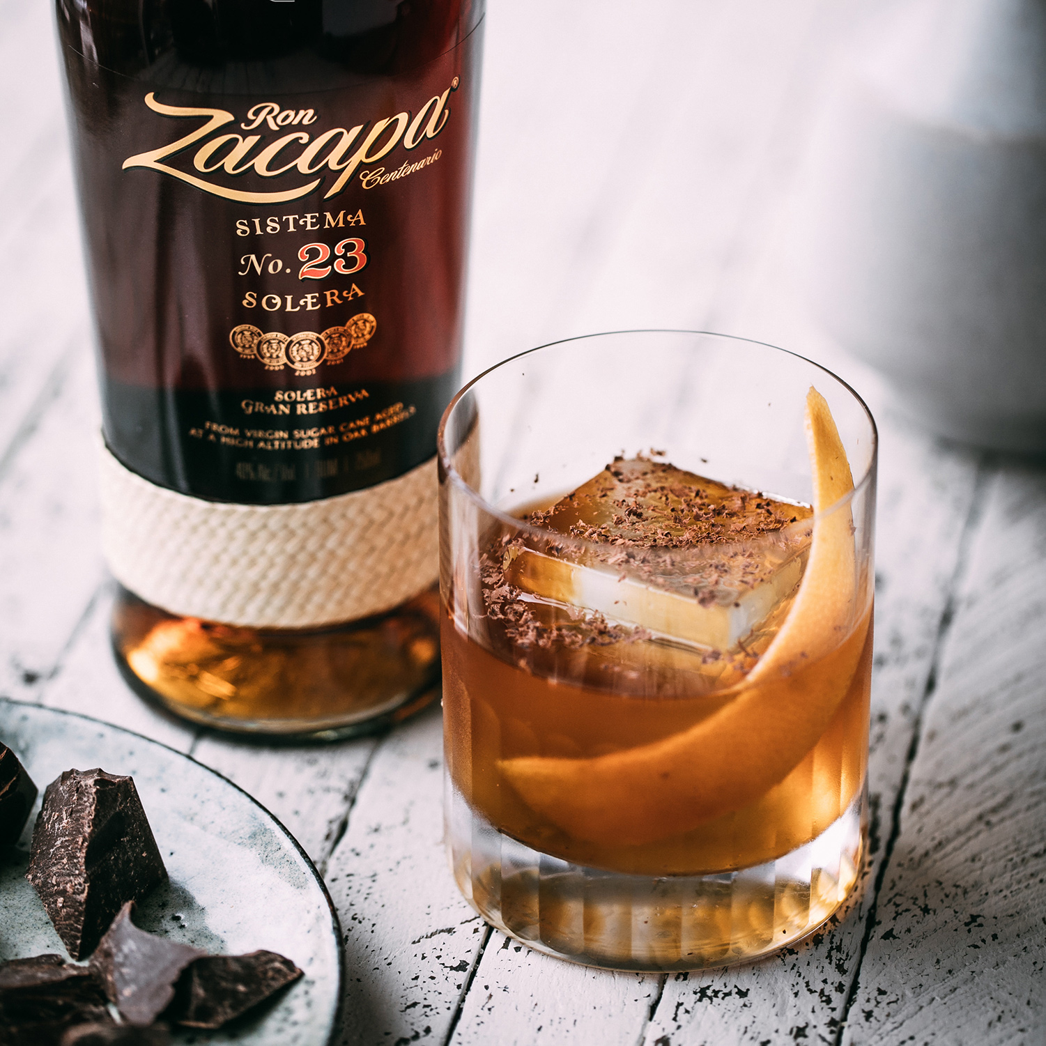 A bottle of Ron Zacapa Sistema 23 Solera rum is positioned on a rustic white wooden surface next to a glass containing a cocktail.