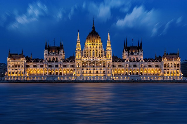 a large building with a gold and black roof and a body of water with Hungarian Parliament Building in the background