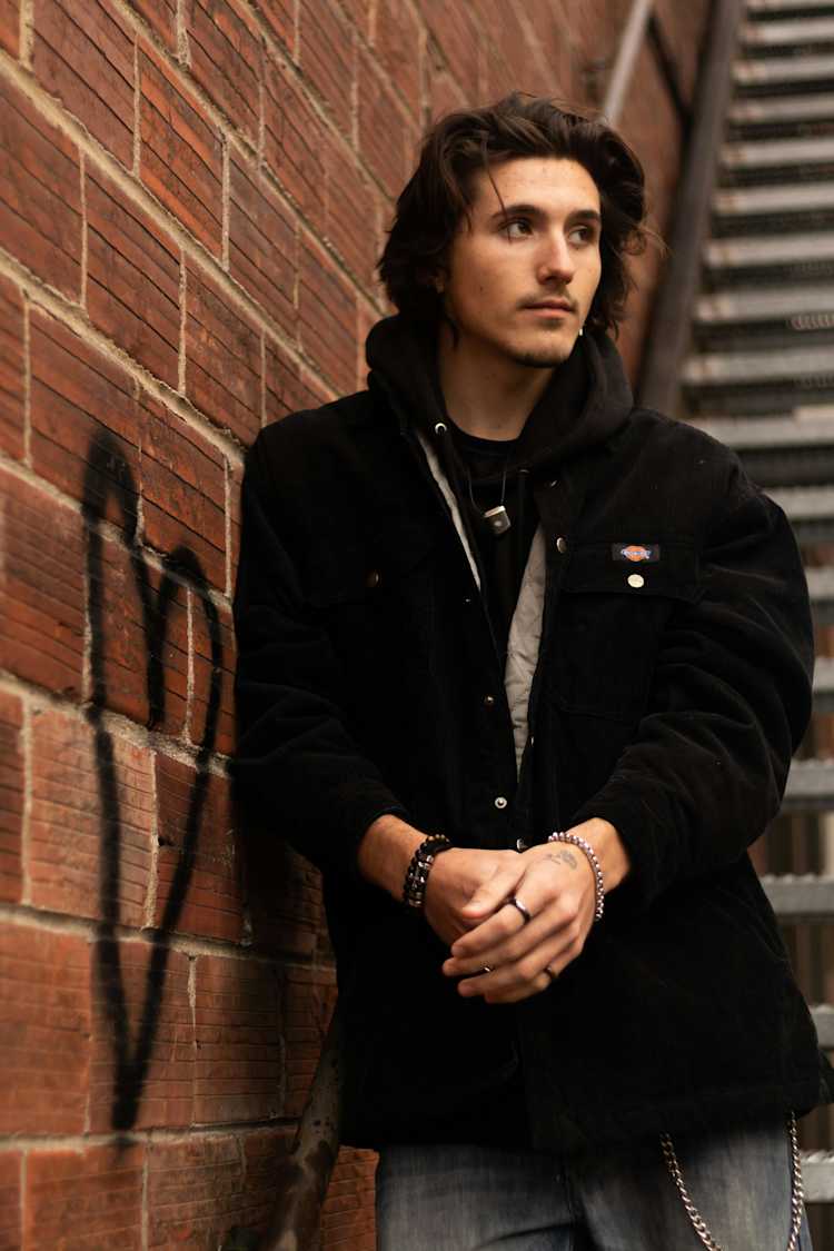 Man in black hoodie and jacket leaning on a brick wall.