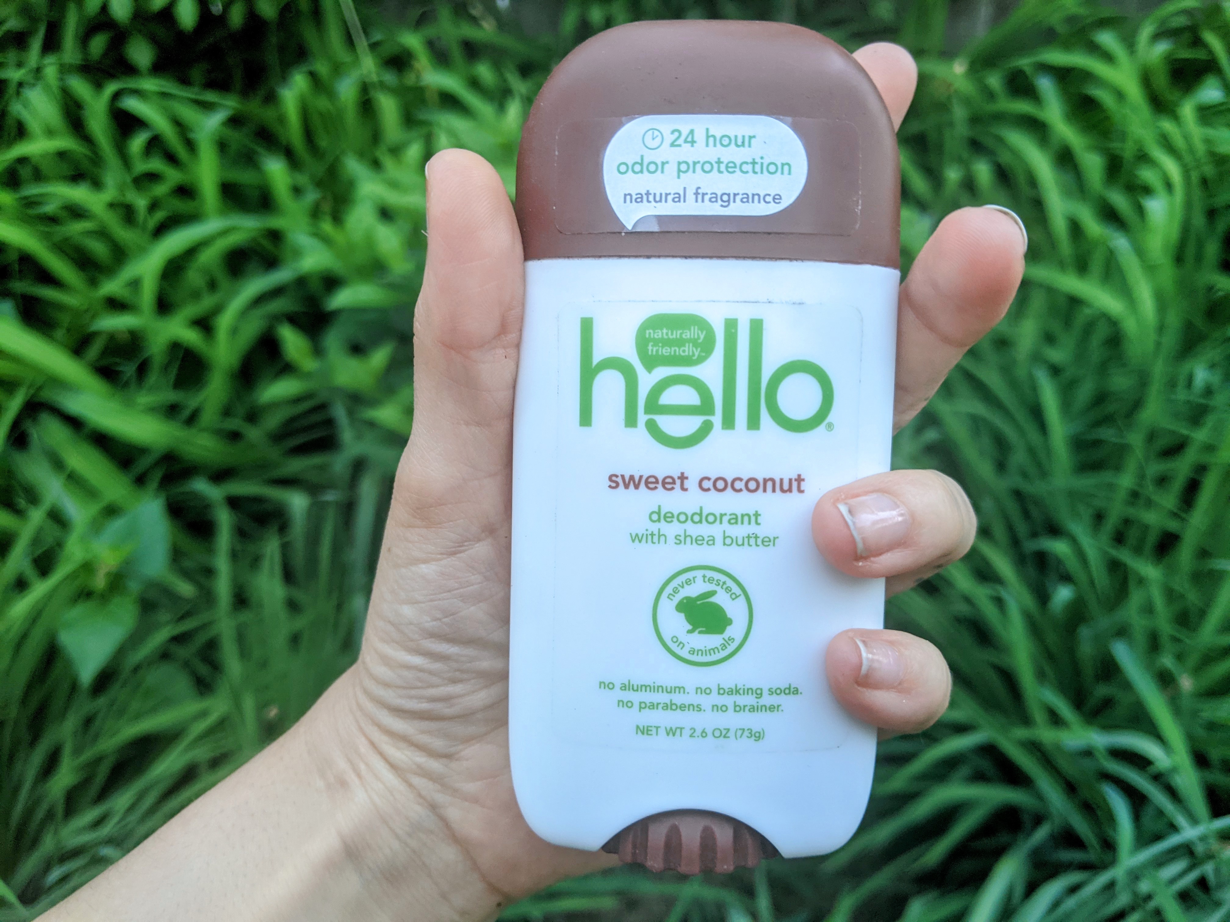 Photo of hand holding up Hello Sweet Coconut deodorant against foliage