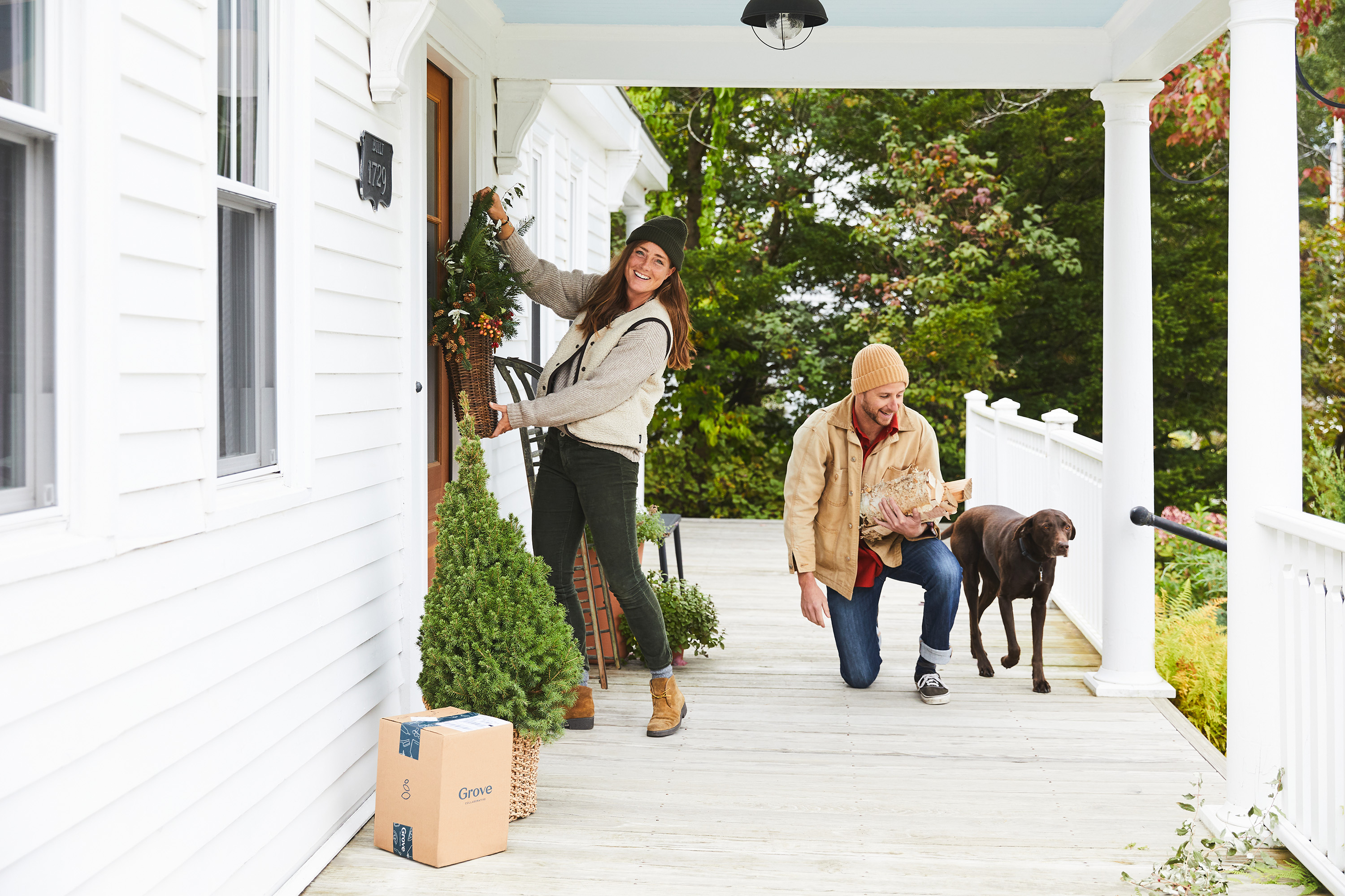 Image of woman hanging wreath on front door while man kneels next to brown dog on front porch