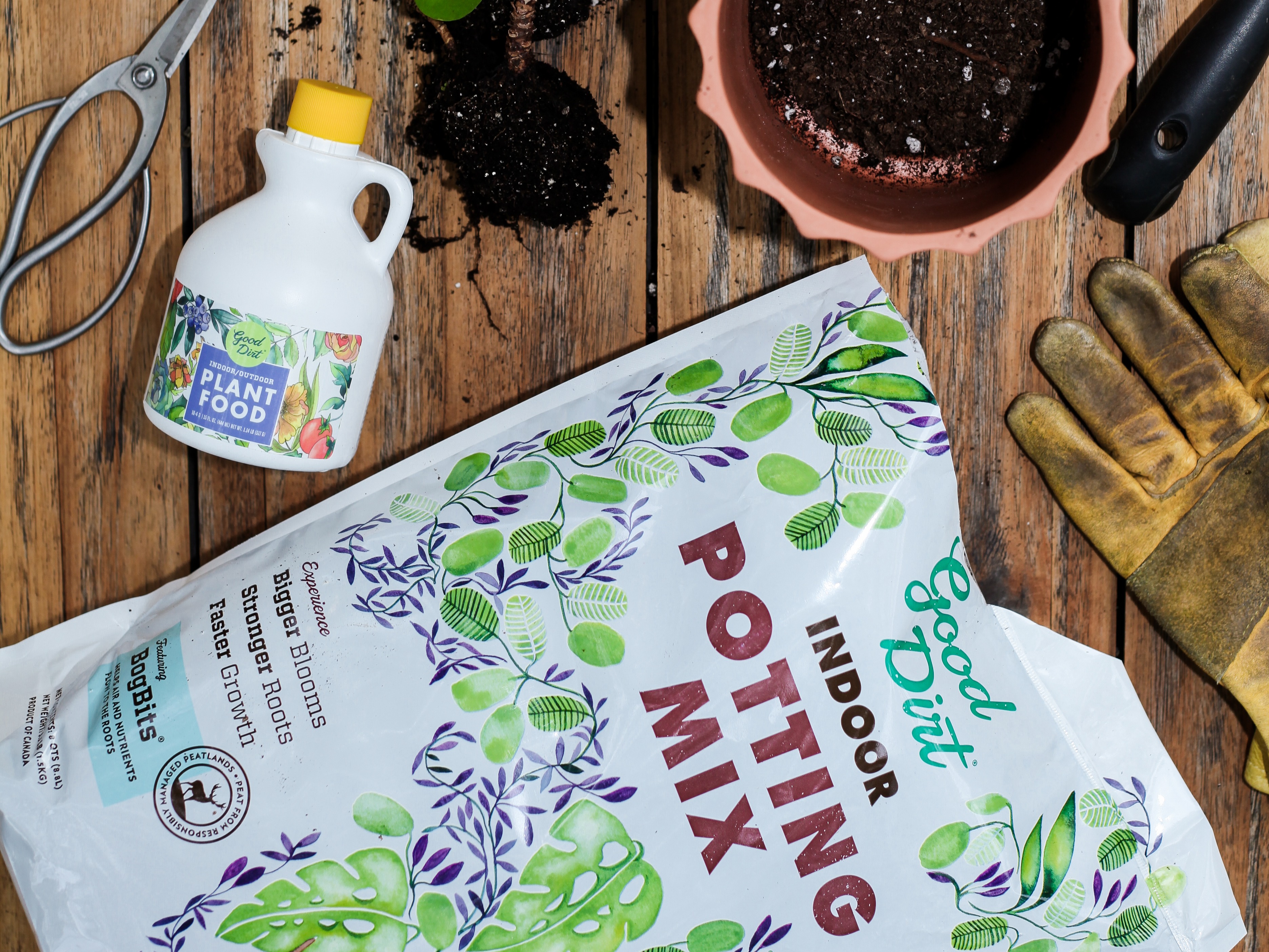 Good Dirt Indoor Potting Mix bag and Good Dirt Plant food on wood deck with gardening gloves and pots