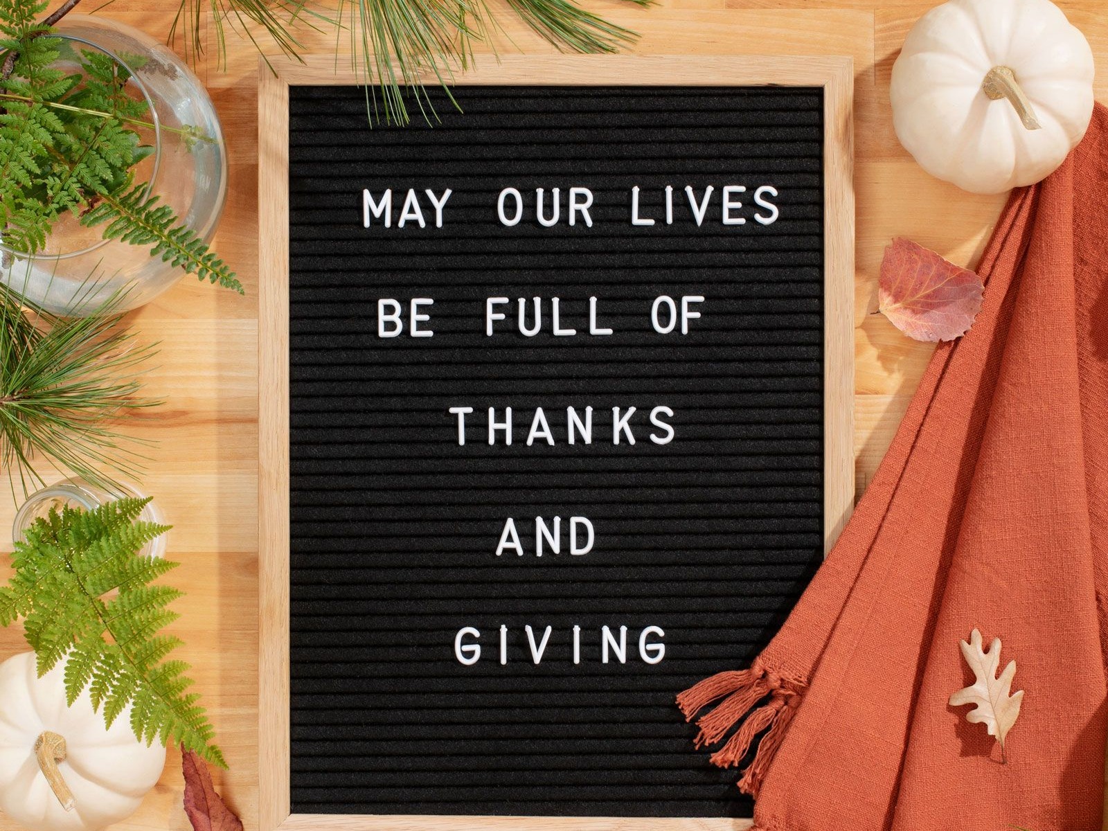 Image of board with white letters that say "May our lives be full of thanks and giving" next to white decorative pumpkin and orange towel