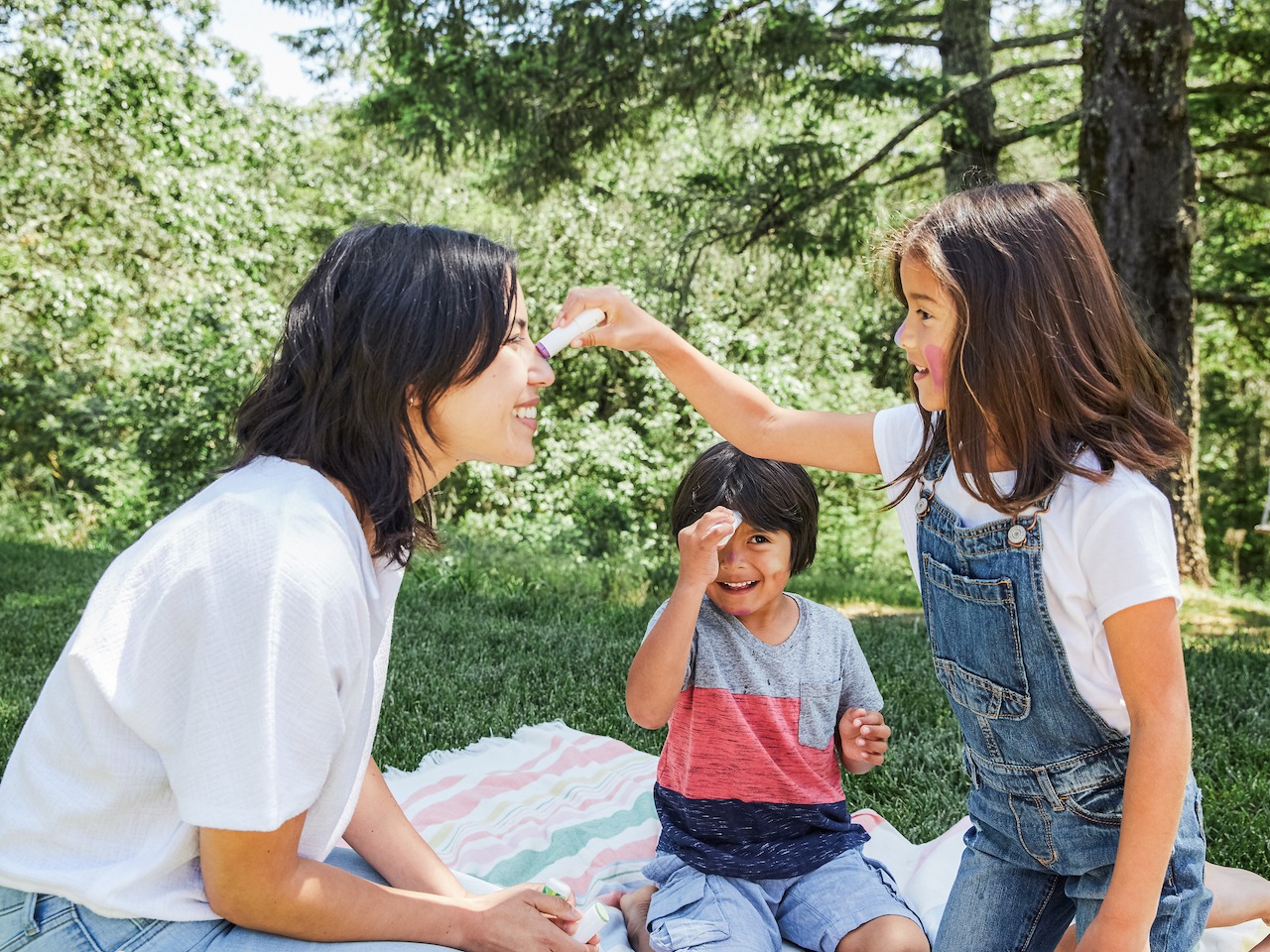Young girl applying sunscreen to a woman outside while a younger boy applies sunscreen on his forehead sitting on a picnic blanket 