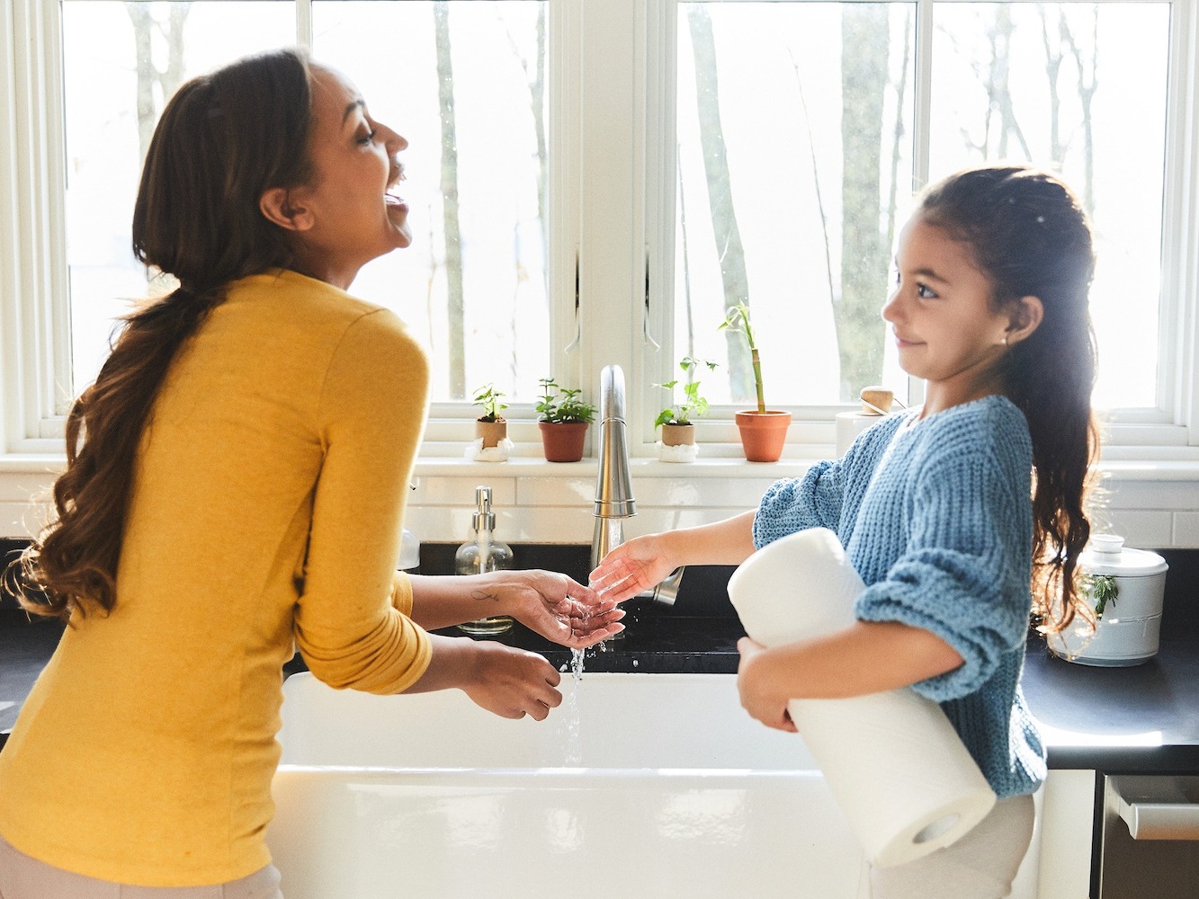 mom and daughter washing their hands at kitchen sink while daughter holds roll of paper towels