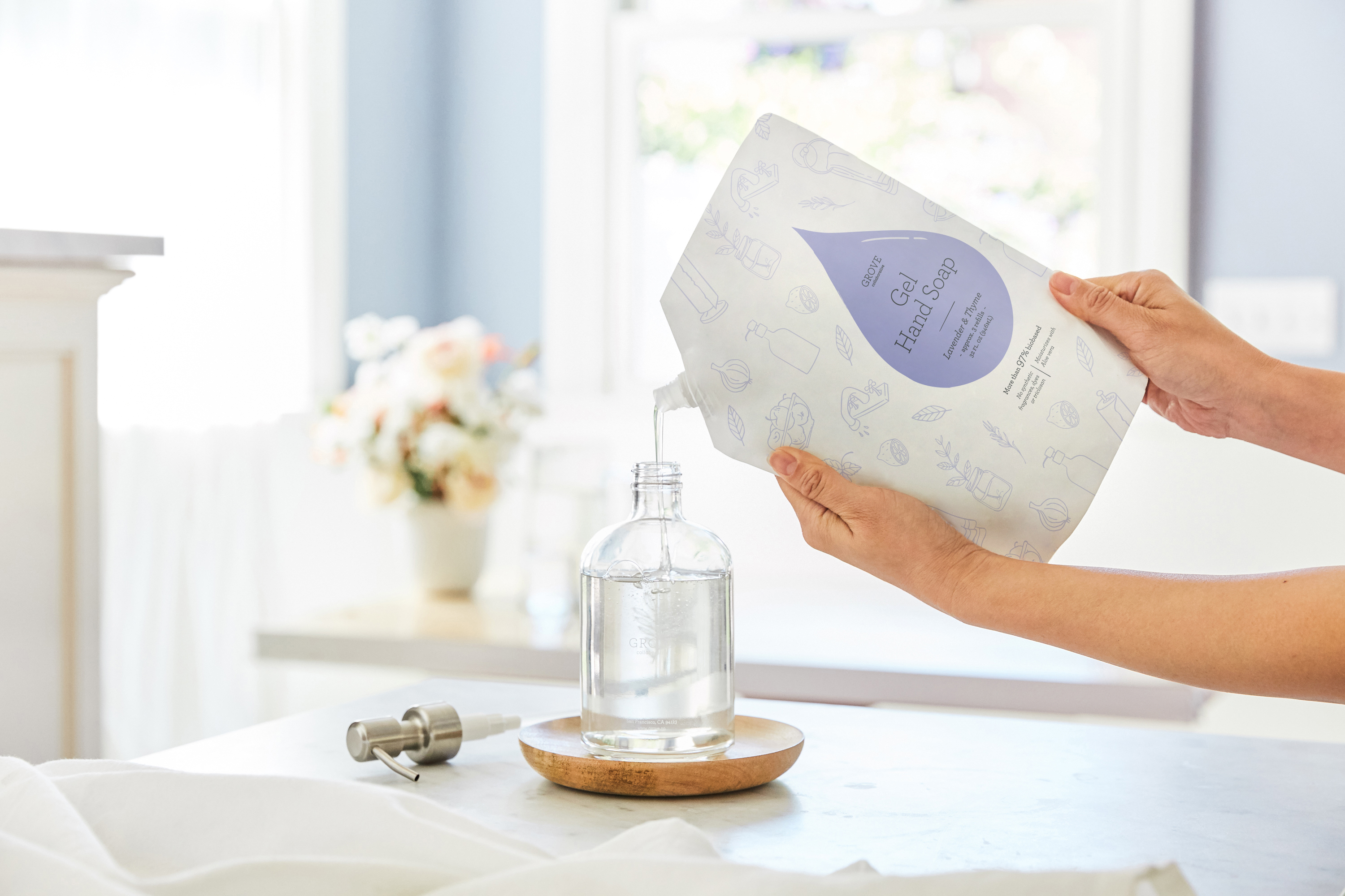 Hands pouring a refill pouch of Grove Gel Hand Soap into a clear soap dispenser
