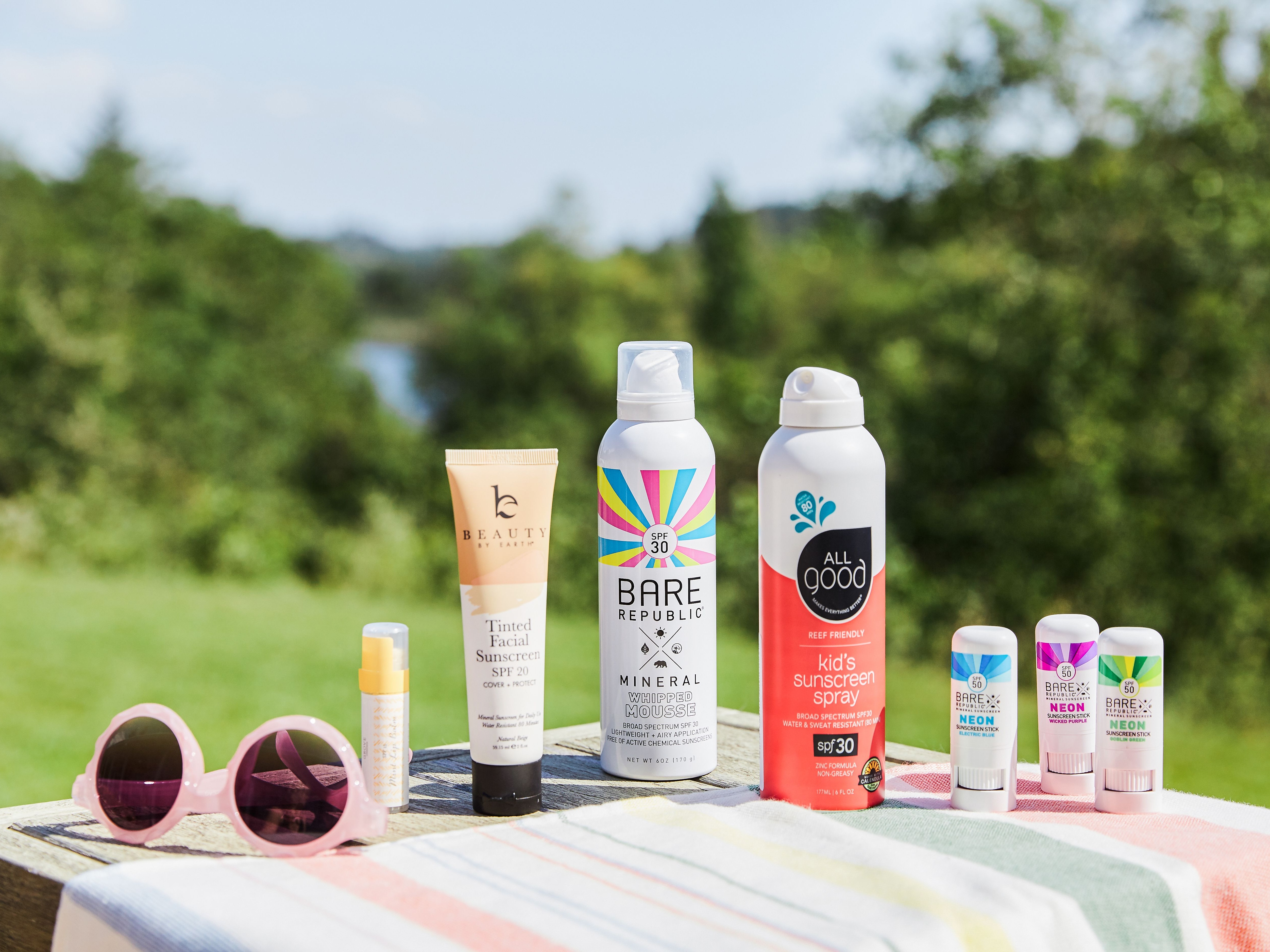 Image of several bottles of brands of mineral sunscreen on striped tablecloth next to pink sunglasses