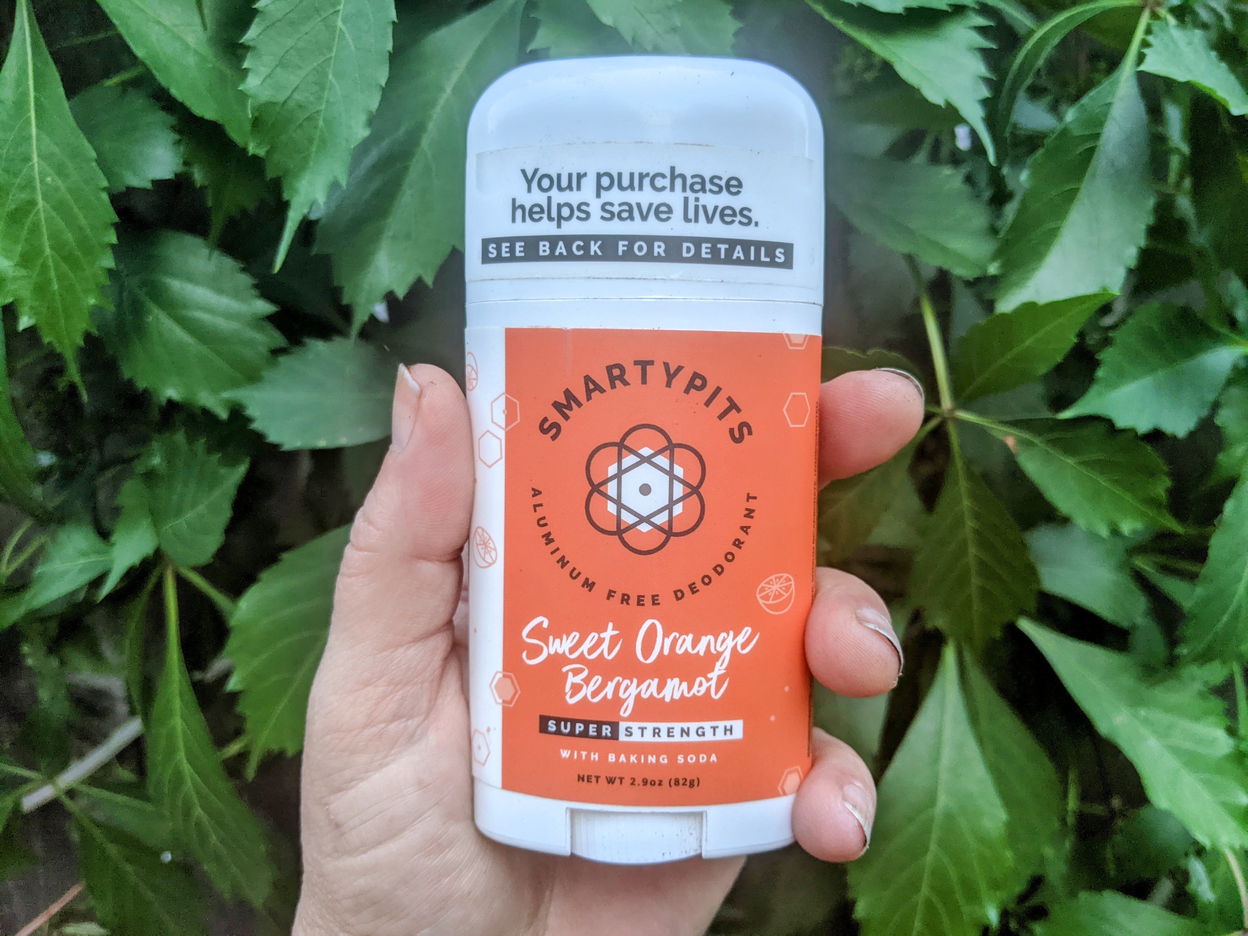 Photo of hand holding up Smarty Pits Sweet Orange Bergamot deodorant against foliage