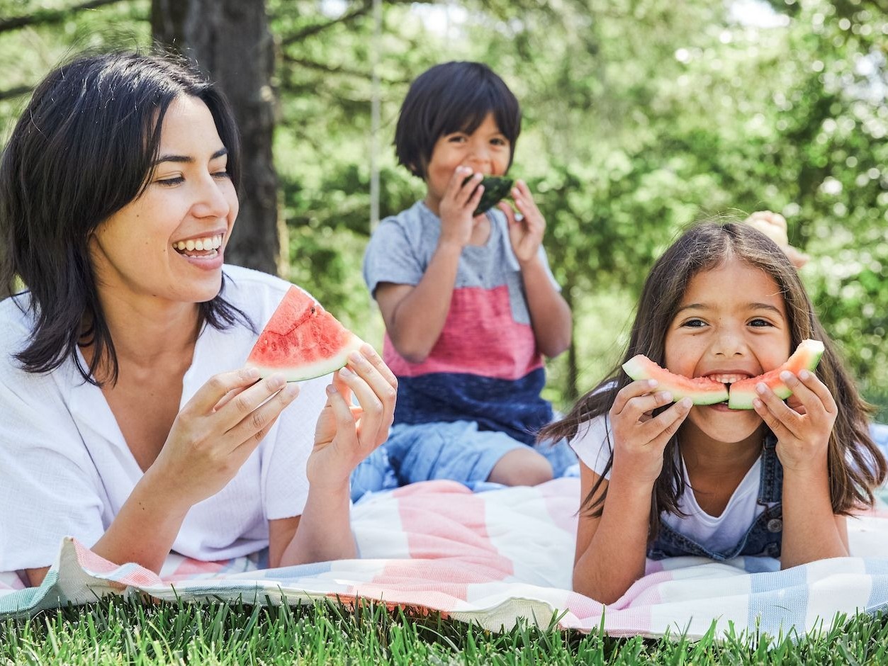 Young woman and young boy and girl laying on a picnic blanket outside in the woods eating watermelon
