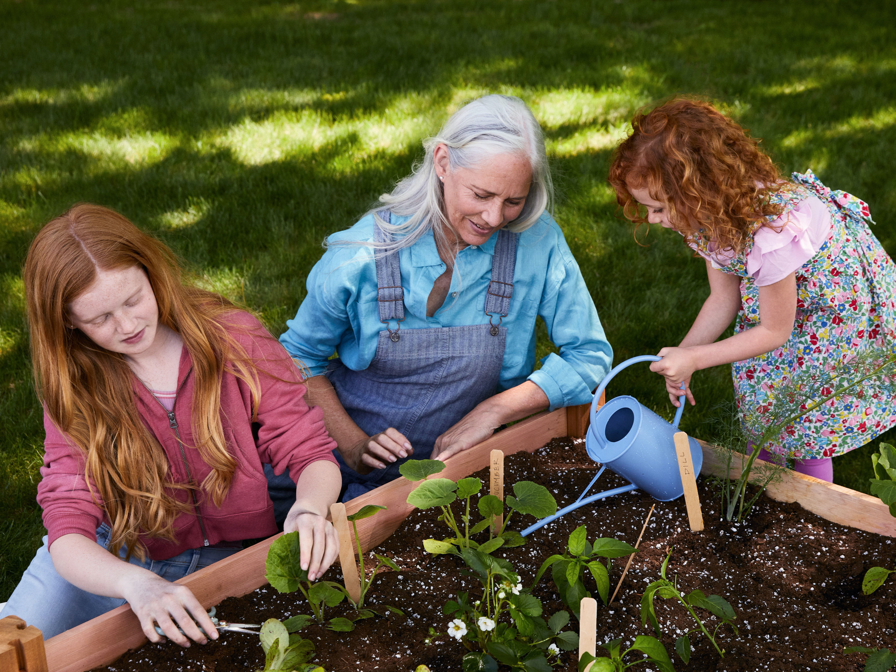 A woman and two young children outside tending to their garden box. One child is clipping herbs while the other waters the soil.