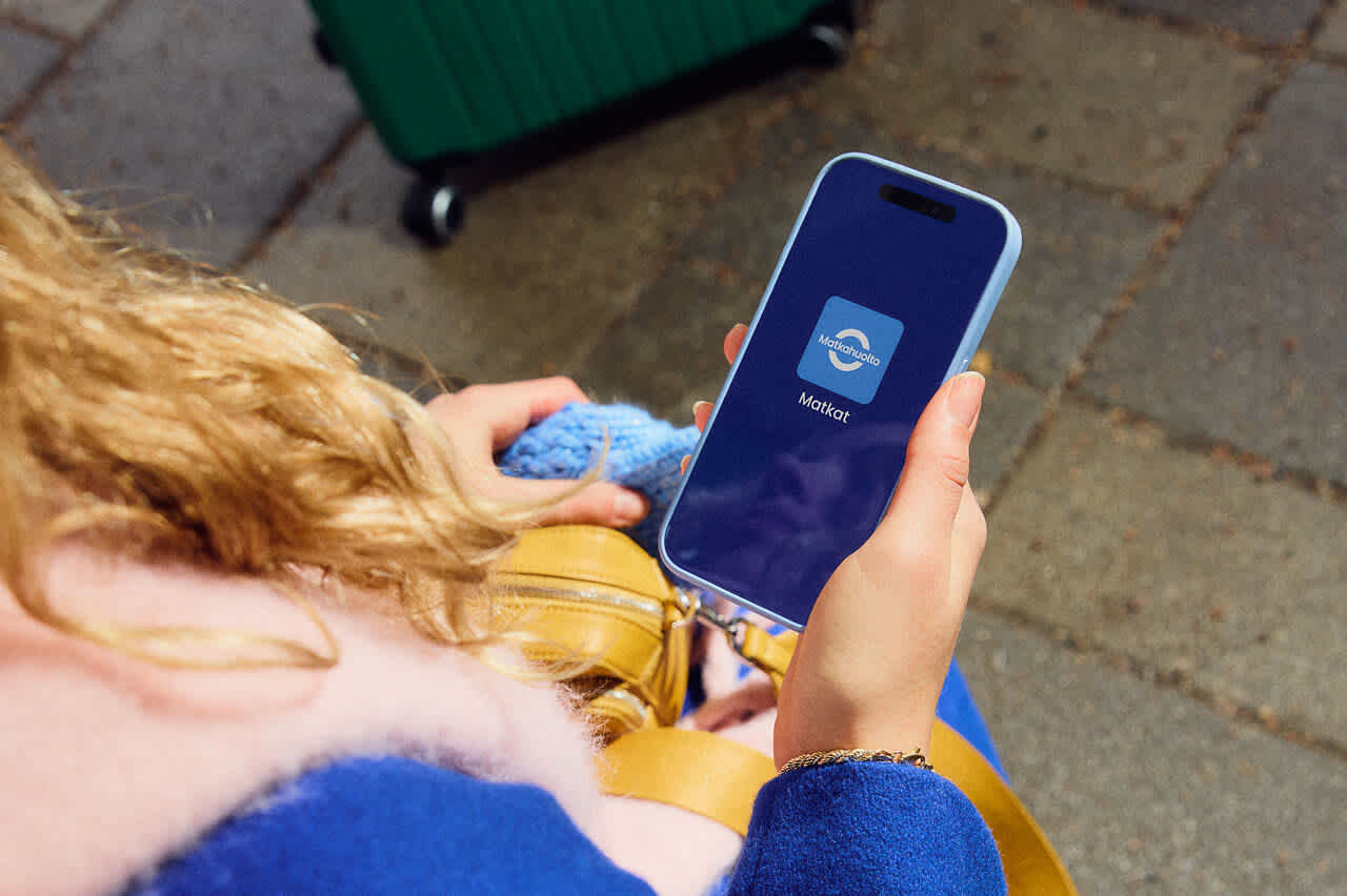 A person looks at the Matkat app icon on a mobile phone screen. The image shows a hand holding a phone and part of a suitcase.