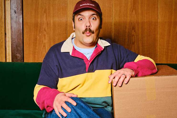 A man sitting on a couch with a cardboard box, making a playful surprised expression against a wooden wall background.