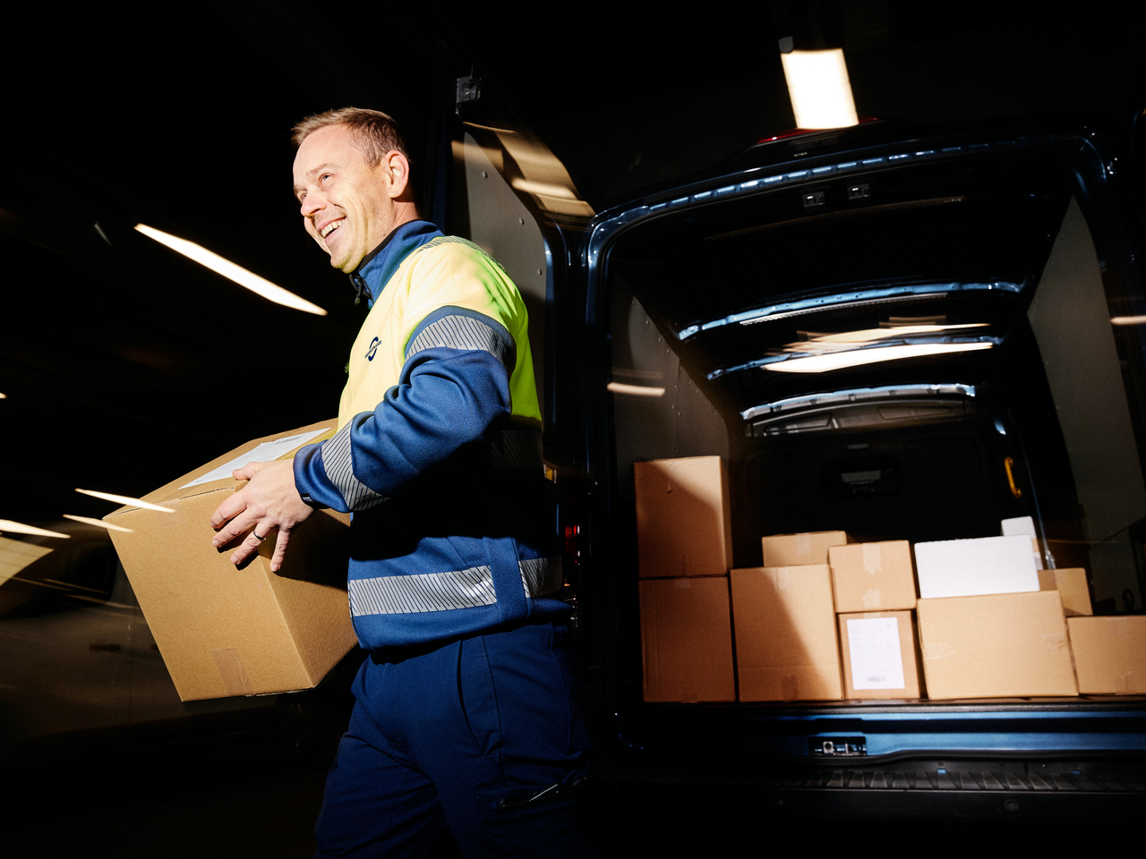 Delivery driver carrying a cardboard box from the open cargo area of a van filled with parcels.