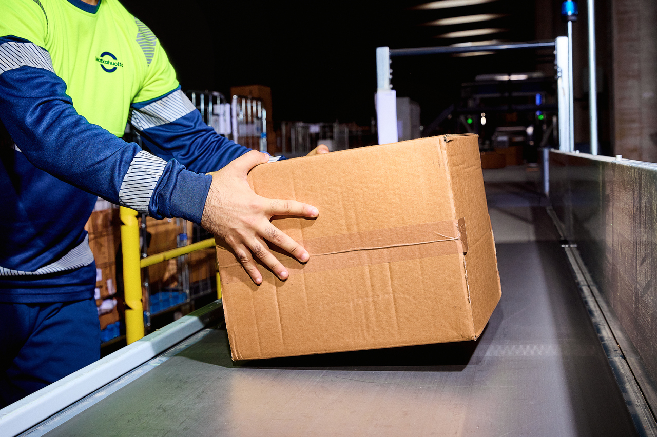 A Matkahuolto employee places a cardboard box onto a conveyor belt at a parcel sorting facility.