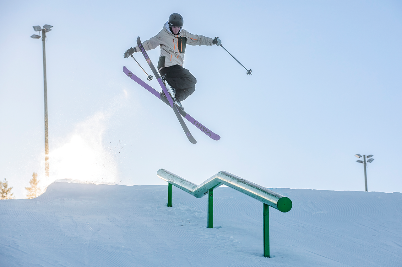 En freestyleåkare utför ett imponerande hopp över en rail i en snötäckt skidbacke under klar vinterhimmel.