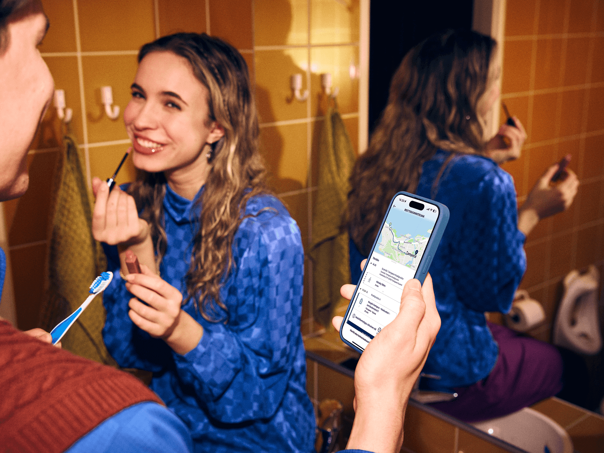 Kaksi henkilöä kylpyhuoneessa; toinen harjaa hampaita ja toinen laittaa huulipunaa peilin edessä. Toisen henkilön kädessä on älypuhelin, jonka näytöllä näkyy Matkahuollon Reitit ja Liput -sovellus ja matkareitti.