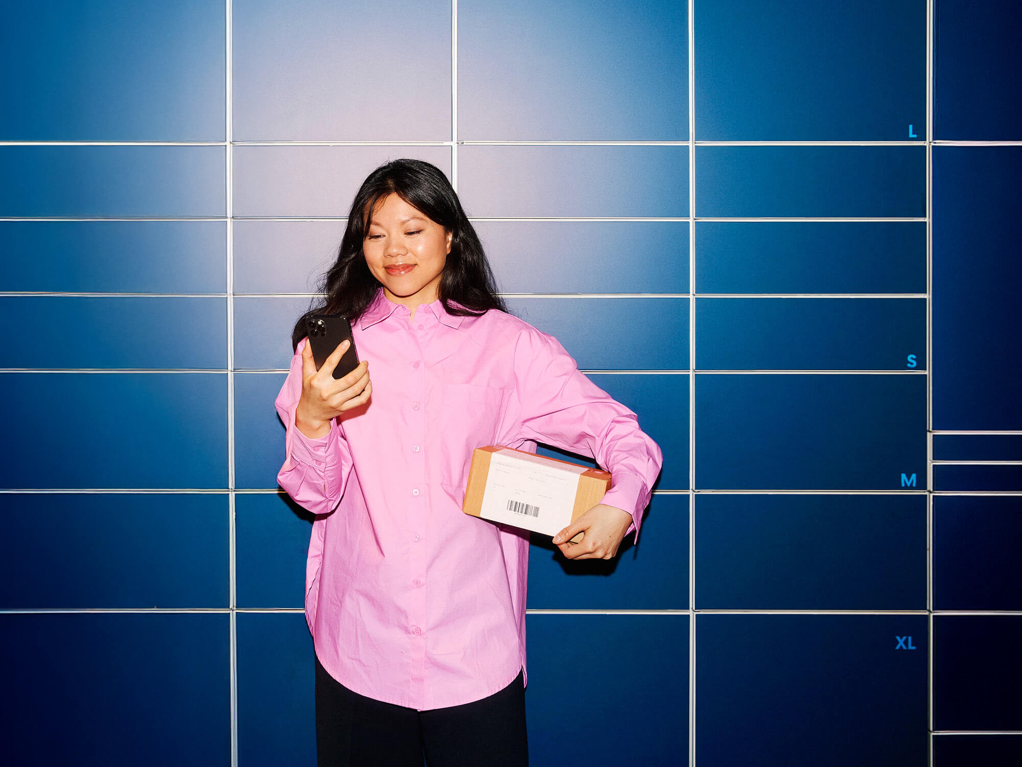 Nuori nainen katsoo puhelintaan ja pitelee kädessään pientä pakettia pakettiautomaatin edessä.