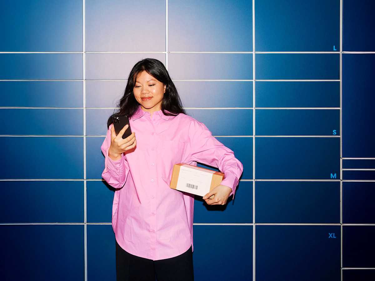 Nuori nainen katsoo puhelintaan ja pitelee kädessään pientä pakettia pakettiautomaatin edessä.