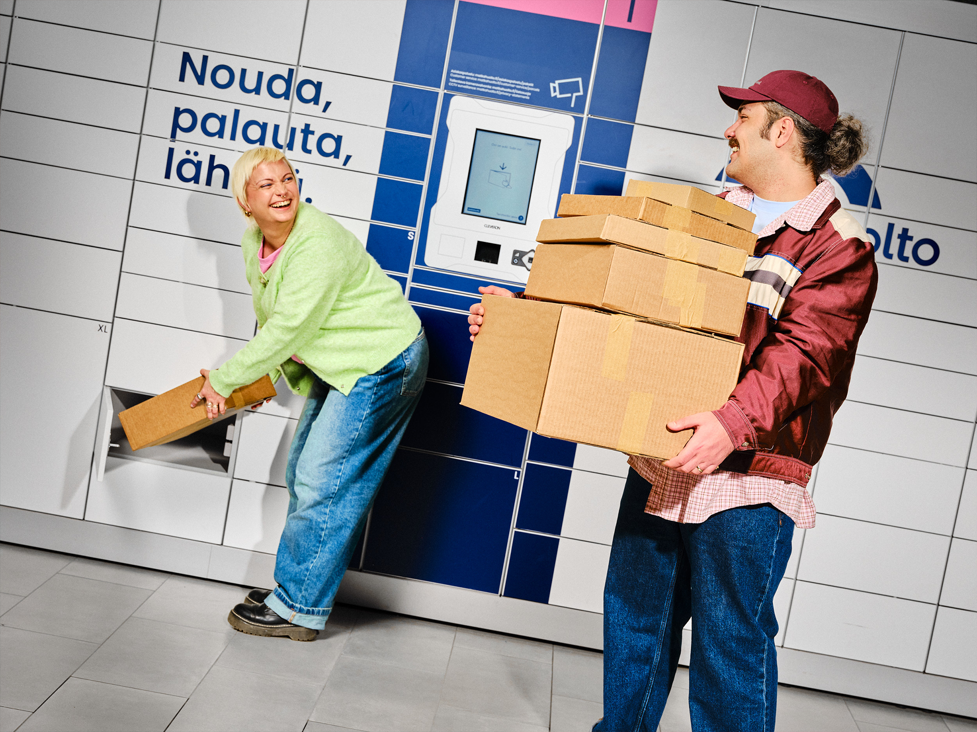 A woman inserts a parcel into a locker while a man holds multiple cardboard boxes in front of a Matkahuolto parcel locker, both smiling.