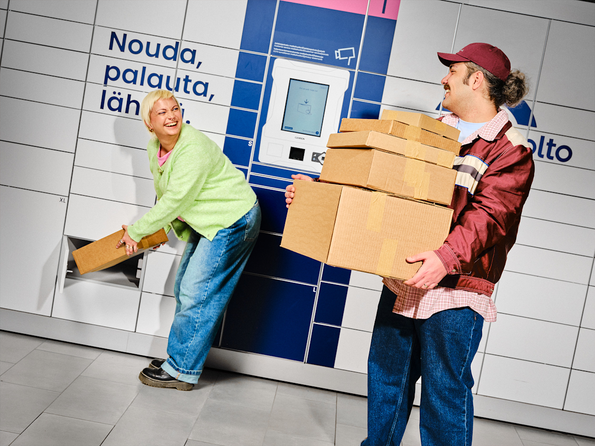 A woman inserts a parcel into a locker while a man holds multiple cardboard boxes in front of a Matkahuolto parcel locker, both smiling.