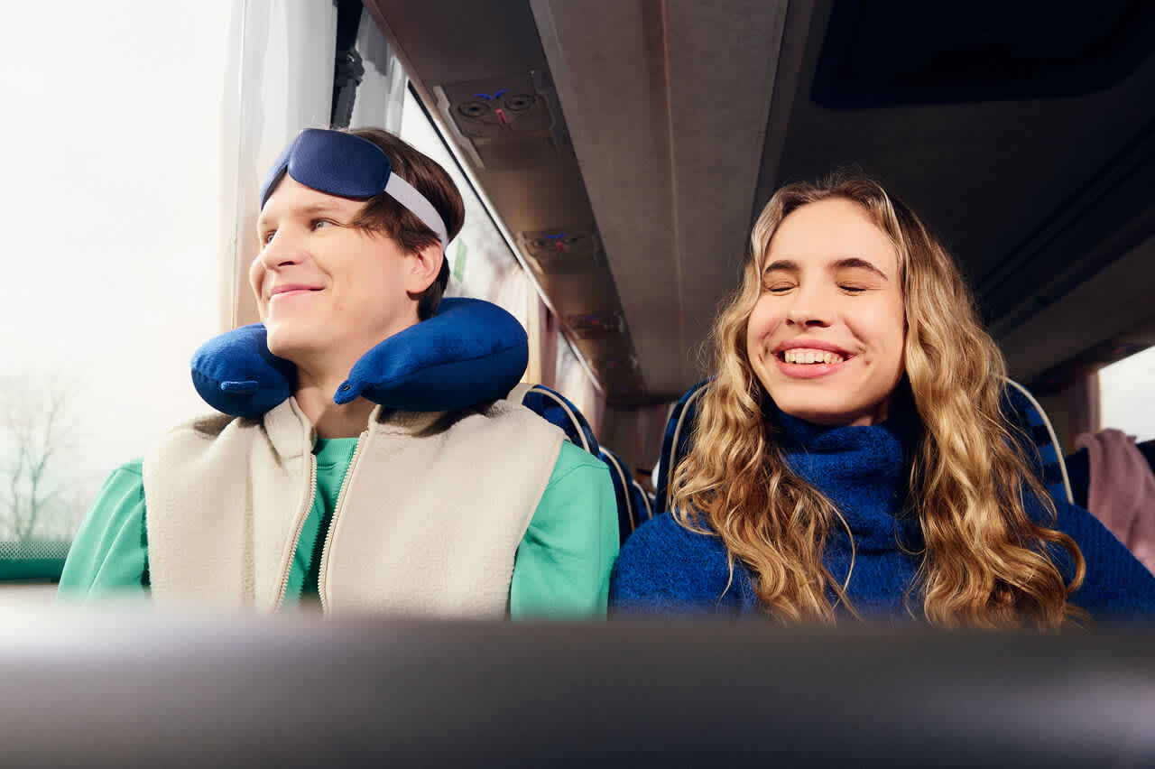 Two passengers are seated on a coach during a journey. One is using a neck pillow and eye mask, while the other is wearing a scarf. The landscape is visible through the window.