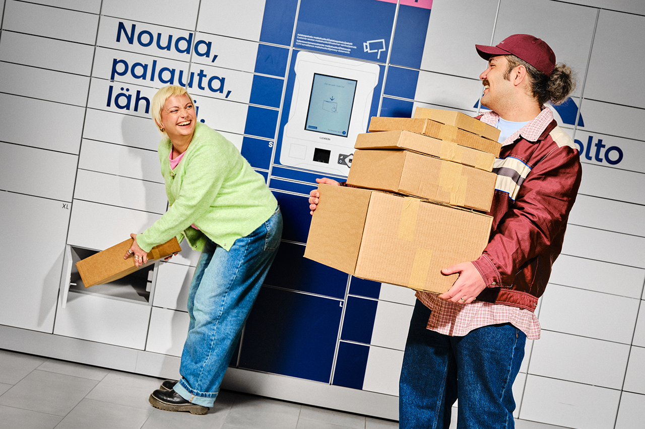 A woman inserts a parcel into a locker while a man holds multiple cardboard boxes in front of a Matkahuolto parcel locker, both smiling.