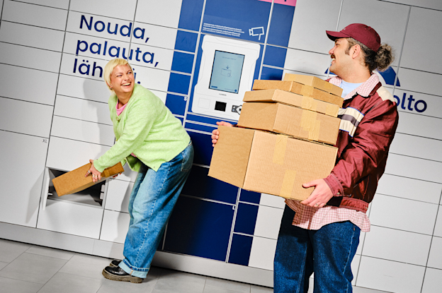 A woman inserts a parcel into a locker while a man holds multiple cardboard boxes in front of a Matkahuolto parcel locker, both smiling.