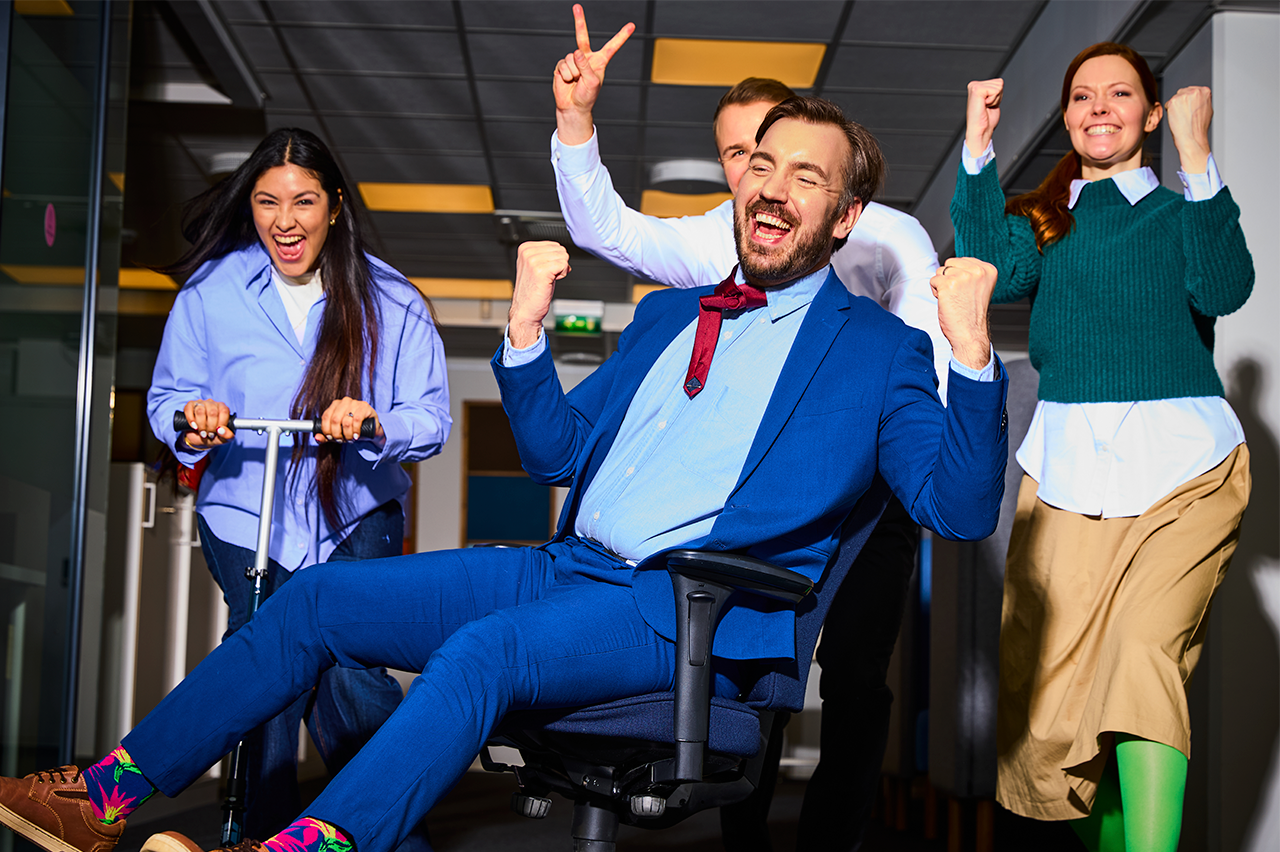 Toimistotiimi juhlii yhdessä – yksi istuu toimistotuolissa muiden työntäessä ja kaikki iloitsevat.