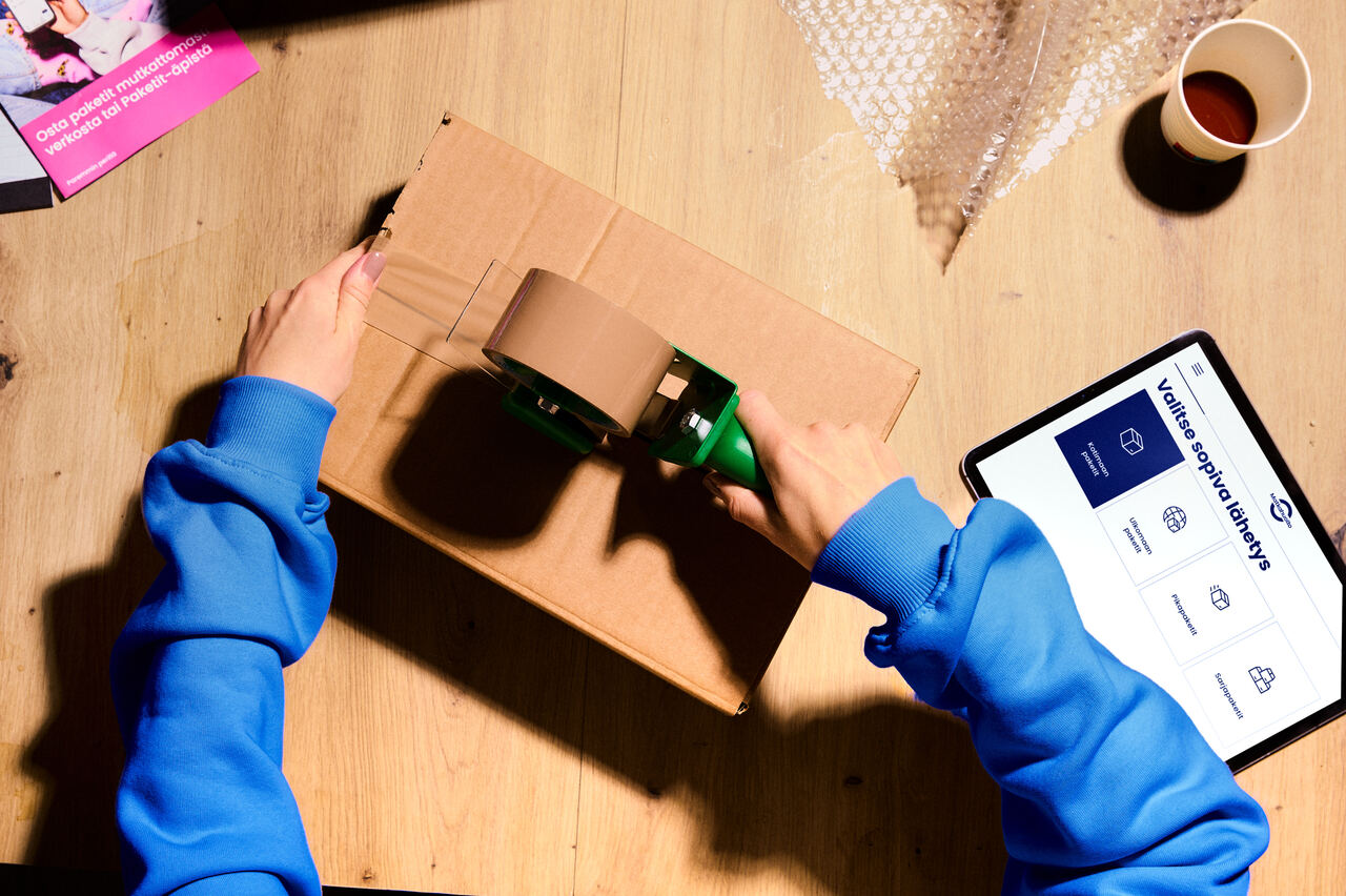 A person seals a cardboard box with packing tape. Next to them, Matkahuolto’s parcel sending service is open on a tablet.