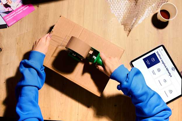 A person seals a cardboard box with packing tape. Next to them, Matkahuolto’s parcel sending service is open on a tablet.
