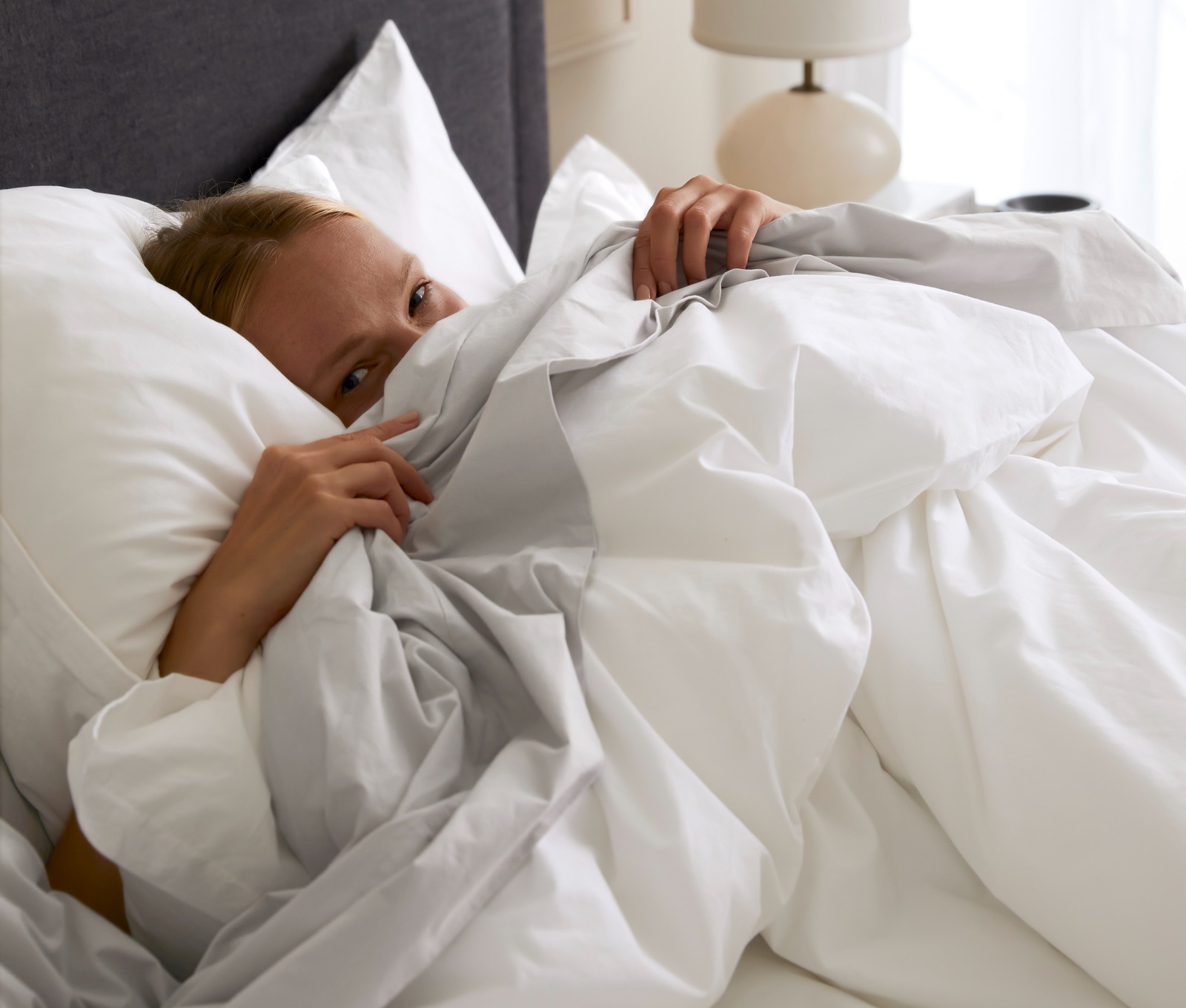 A person peeking out from underneath white and mist brushed cotton sheets