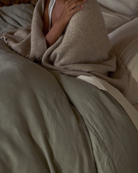 A person sitting in a bed with a cozy blanket wrapped around their shoulders and a moss green linen duvet cover