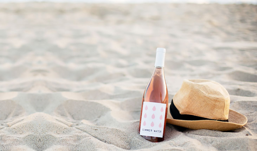 A bottle of rose next to a hat on the beach. 