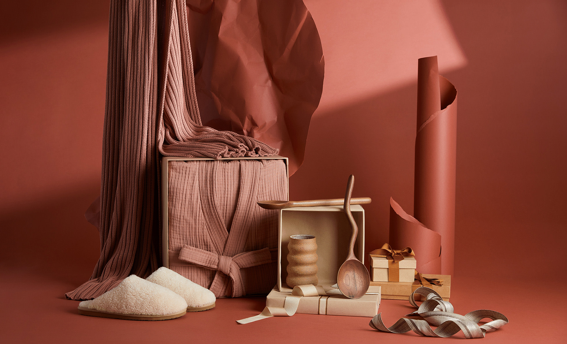An assortment of giftable items: robes, slippers, vases, throw blankets, and decorative wooden spoons against a dusty red background