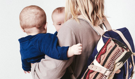 Woman carrying two babies wearing a diaper bag. 