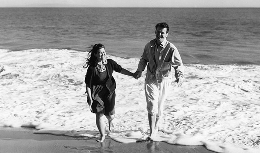 Black and white image of a couple holding hands on the beach. 