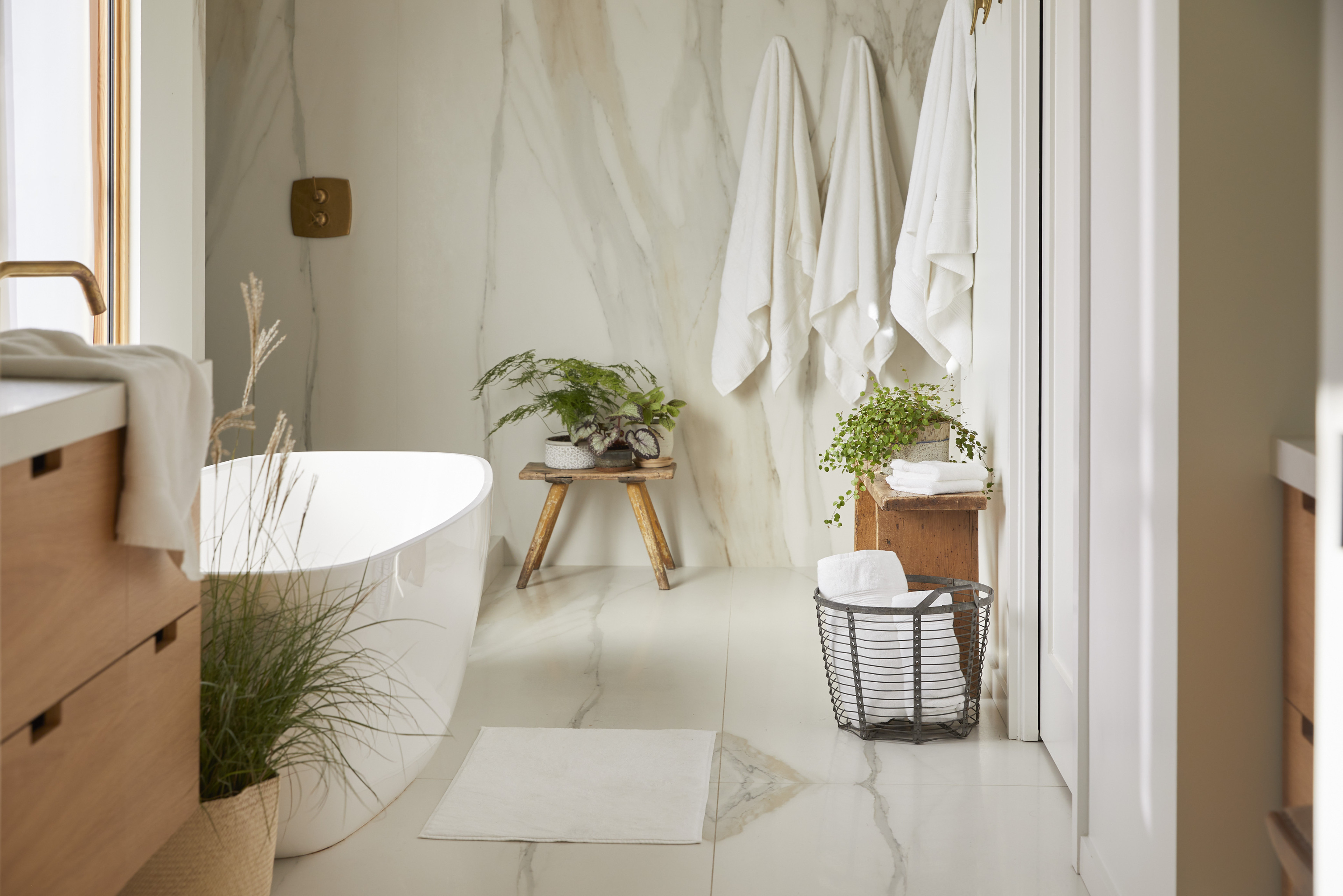 A naturally lit marble bathroom with three white colored robes, several white towels, and one white bathmat.