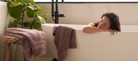 A woman in a modern bathtub with dusty purple towels hanging on the side