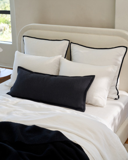 A bed with white sheets with black piping and a decorative pillow