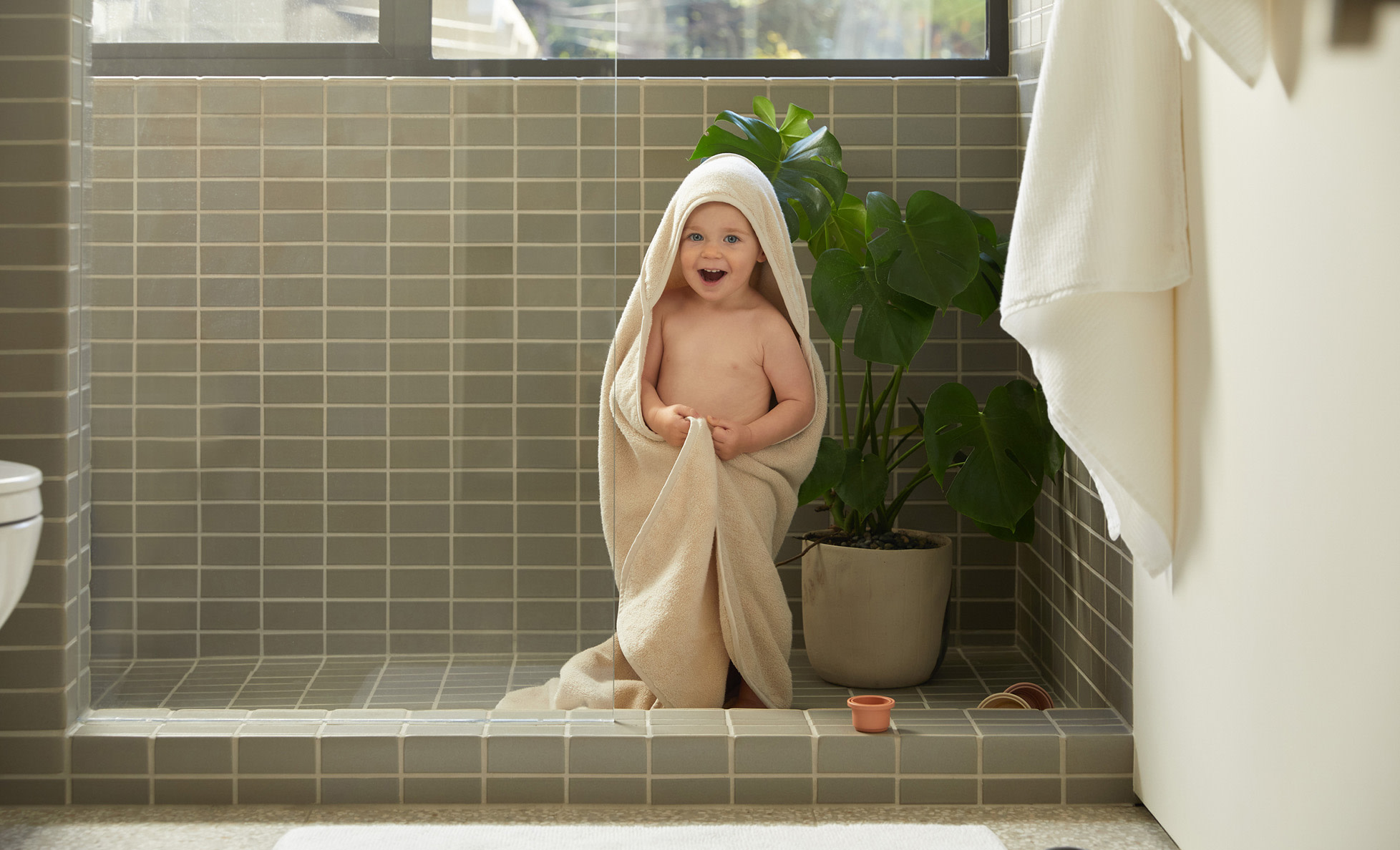 A baby wearing a hooded towel and standing in a green-tiled shower